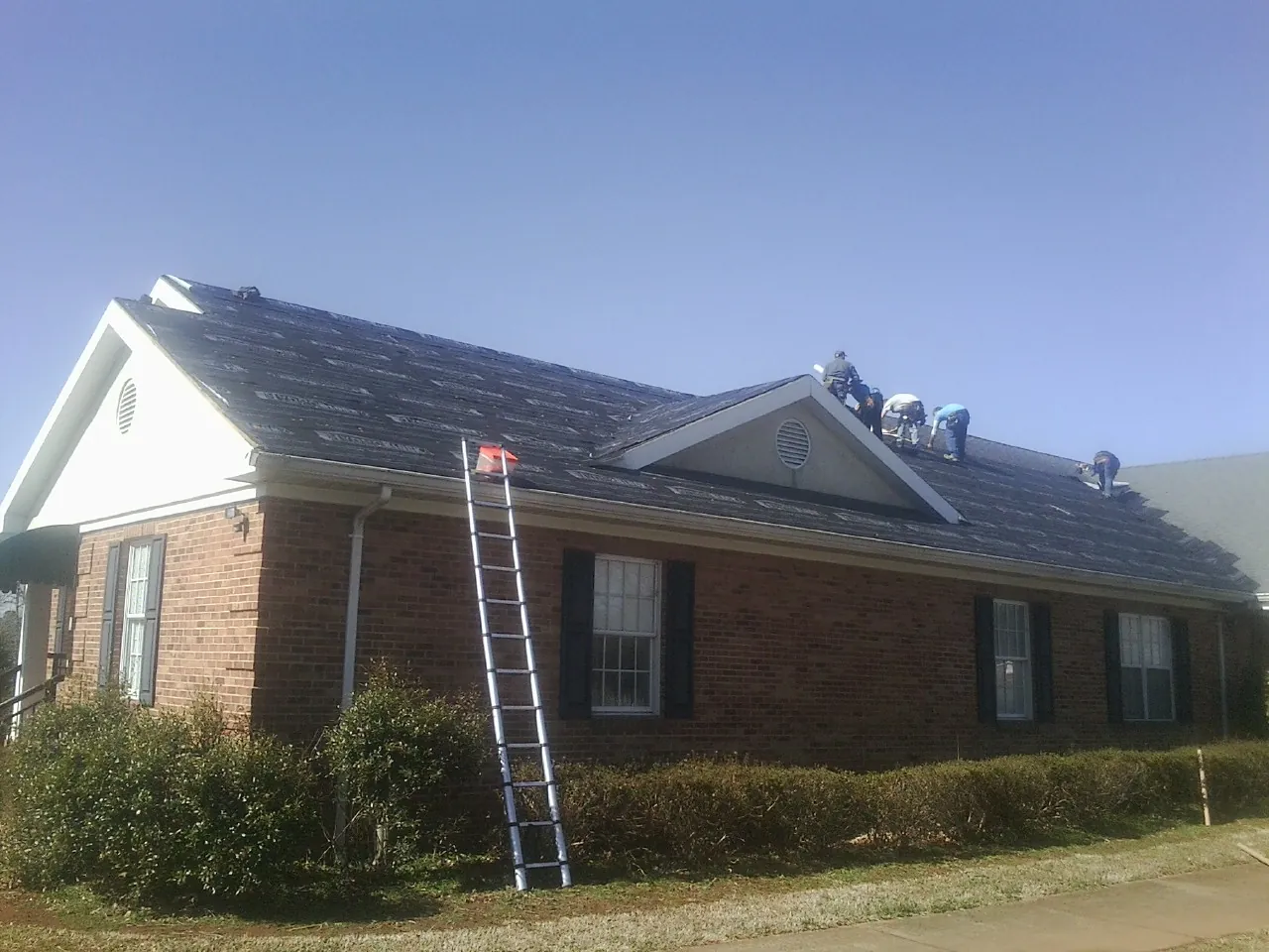 Crew tearing shingles off a brick ranch roof with a ladder set — Emergency Roof Repair work by our crew in Gadsden
