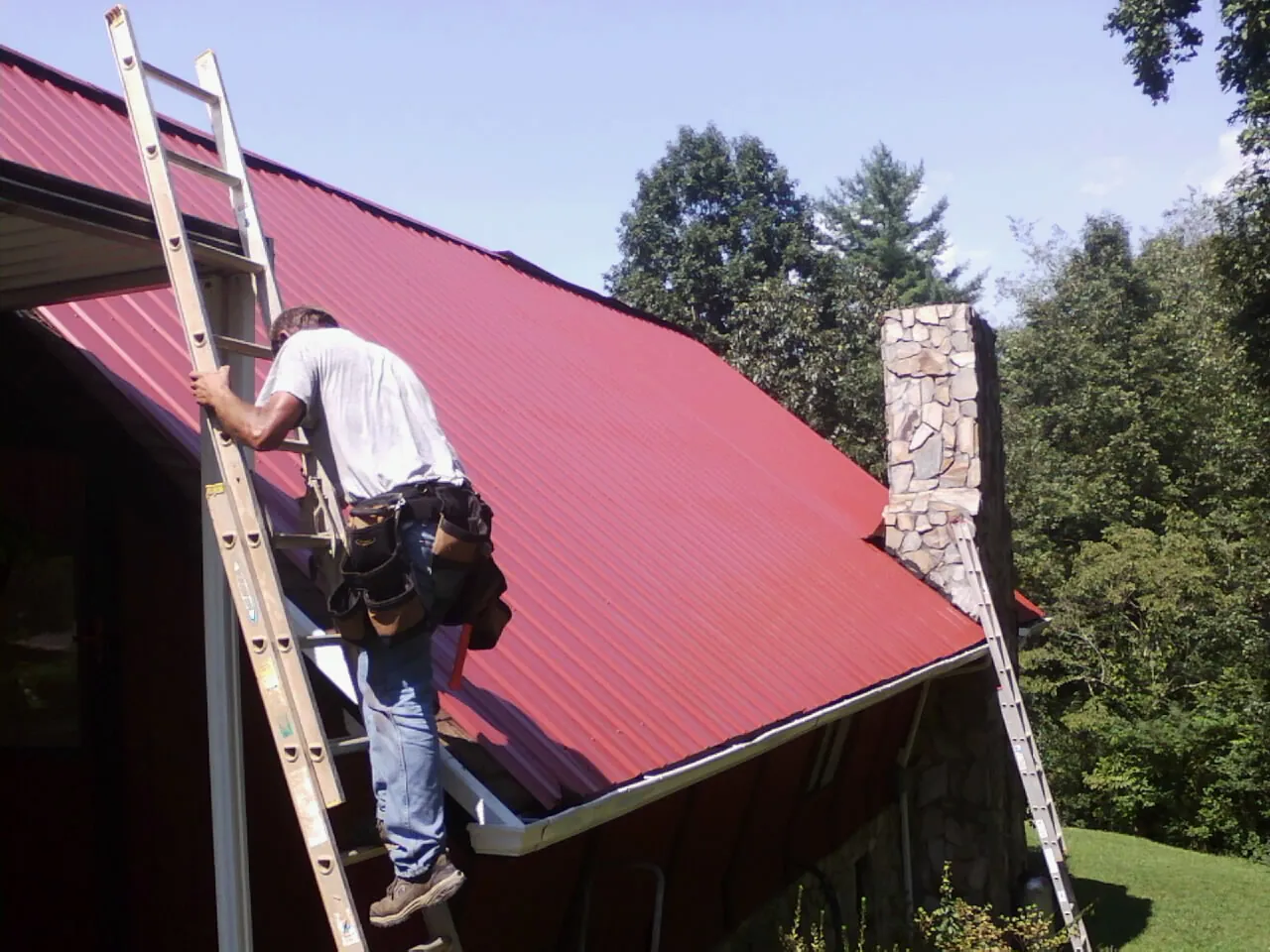 Roofer on a ladder beside a red metal roof and stone chimney — Soffit & Fascia Repair by Roofing Brothers in Gadsden